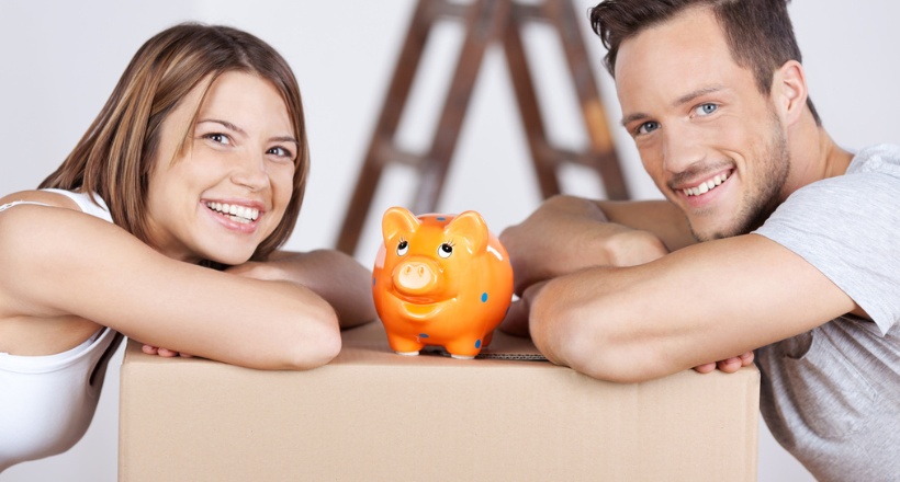 Couple souriant avec une tirelire cochon sur un carton de déménagement, évoquant les économies réalisées grâce à des travaux ciblés